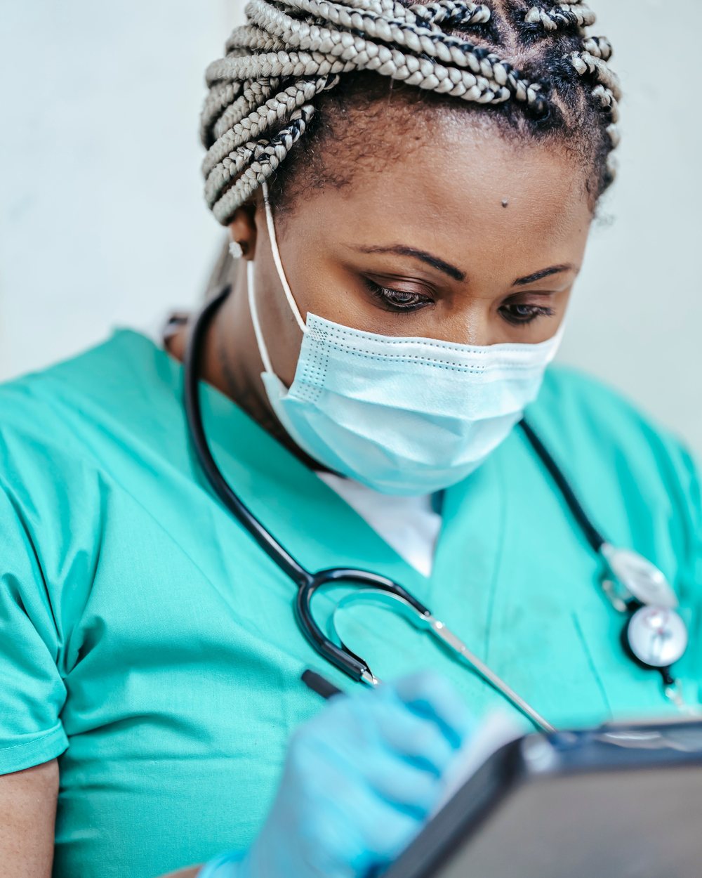 A nurse in scrubs and mask recording notes on a tablet.